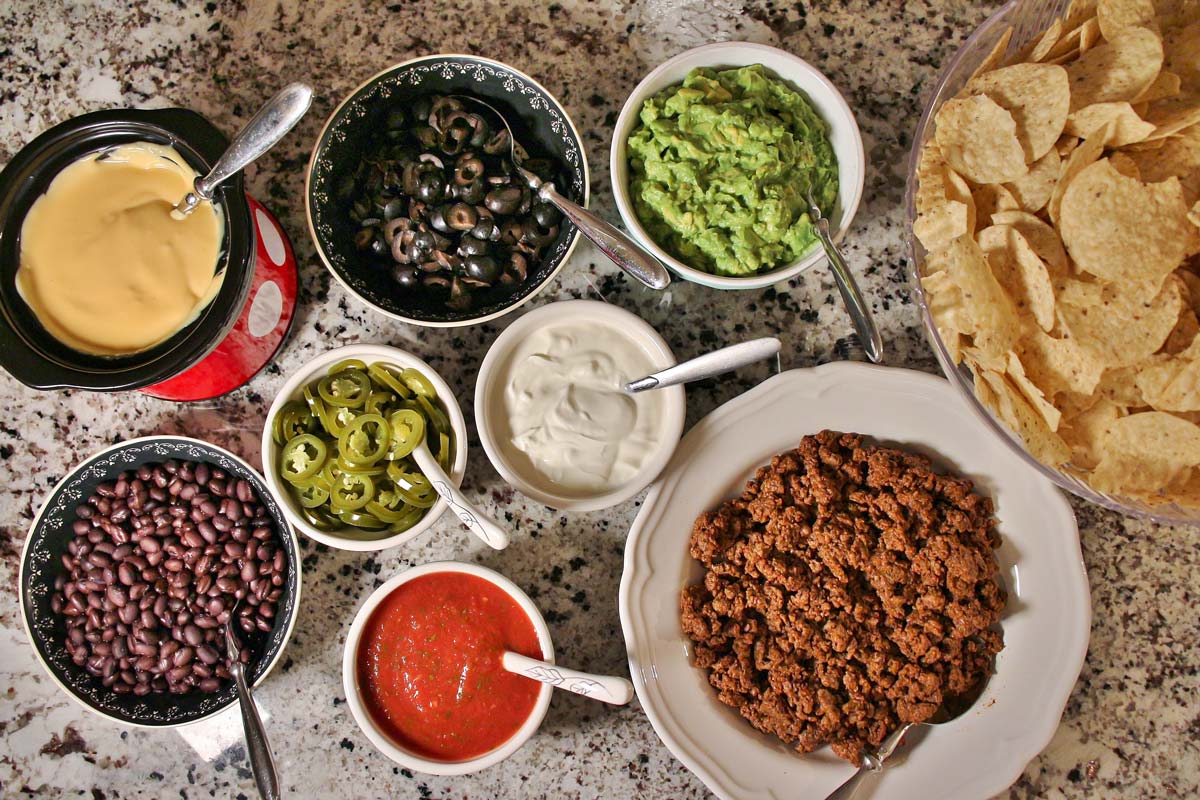 Bowls of tortilla chips, shredded cheese, cooked ground beef, black beans, diced tomatoes, jalapeños, sour cream, and lime arranged on a kitchen counter ready for nachos