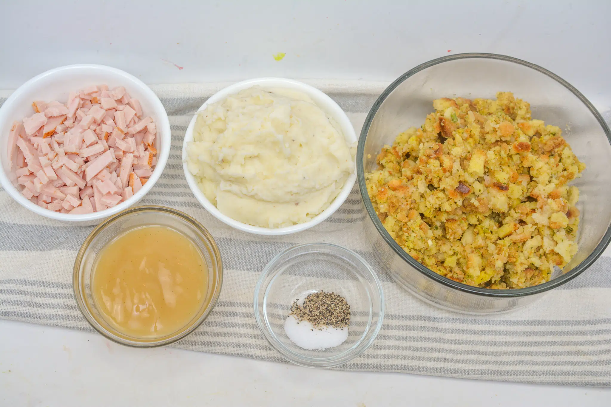 Bread cubes, chopped onion and celery, fresh herbs, eggs, and broth arranged on a clean kitchen counter in warm natural light