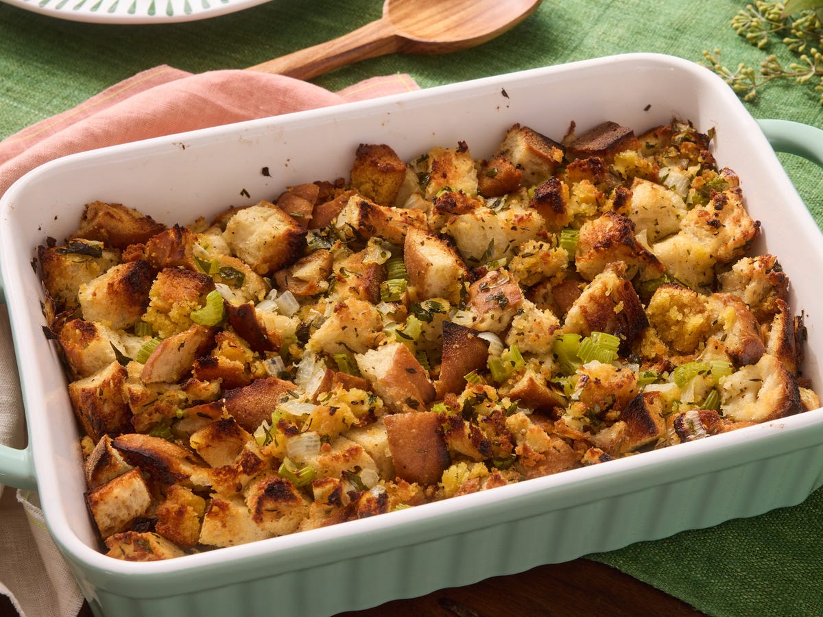 Bread cubes, diced celery, diced onion, fresh herbs, butter, and a carton of broth arranged on a home kitchen counter in warm natural light