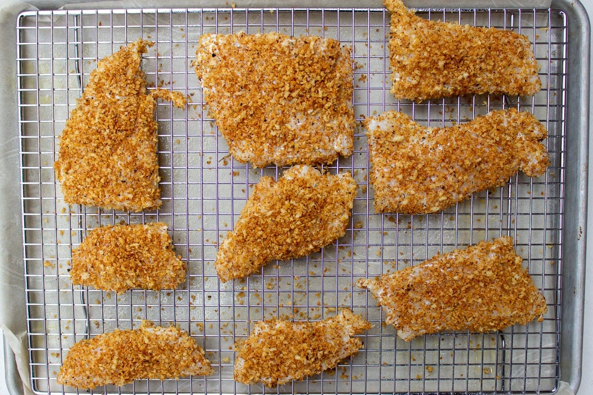 Breaded fish fillets resting on a wire rack before frying, with a skillet heating on the stove in the background