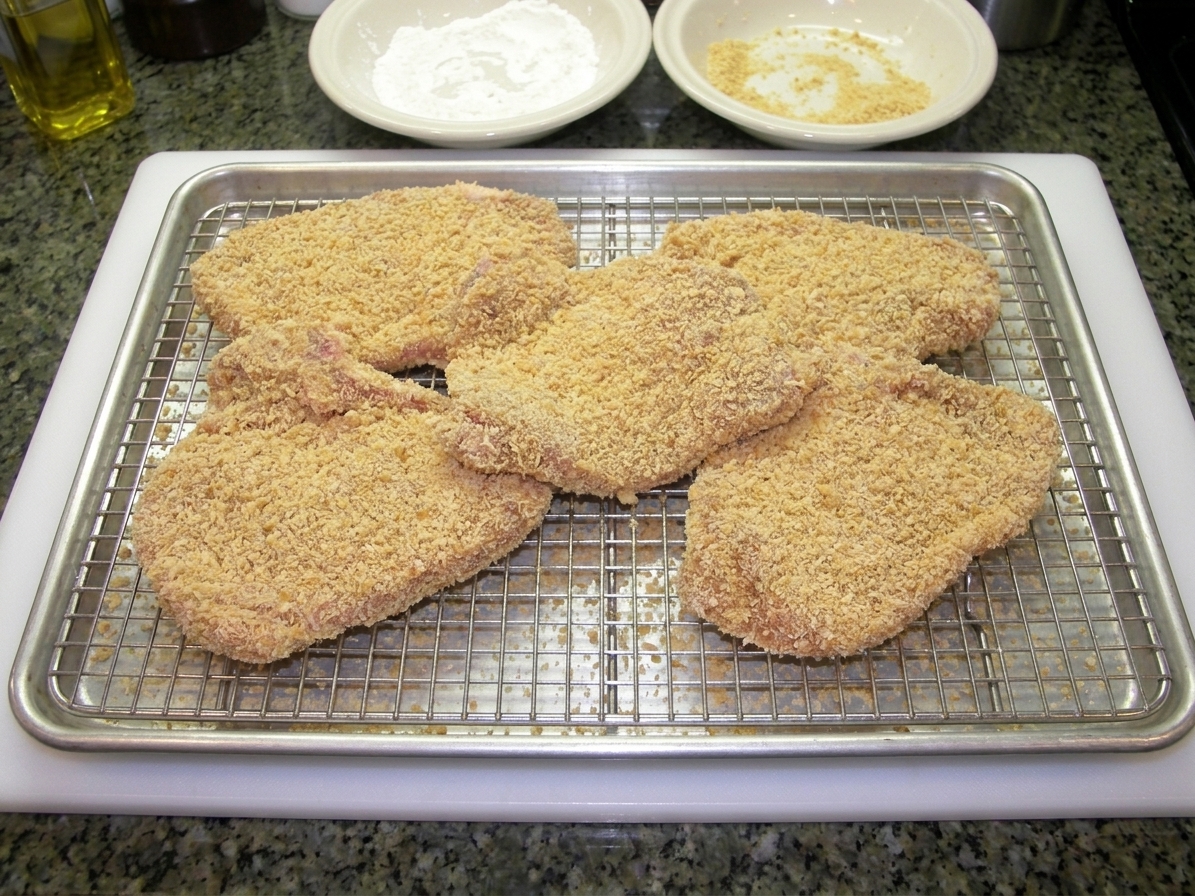 Breaded veal cutlets resting on a wire rack over a sheet pan, ready to fry, in a home kitchen