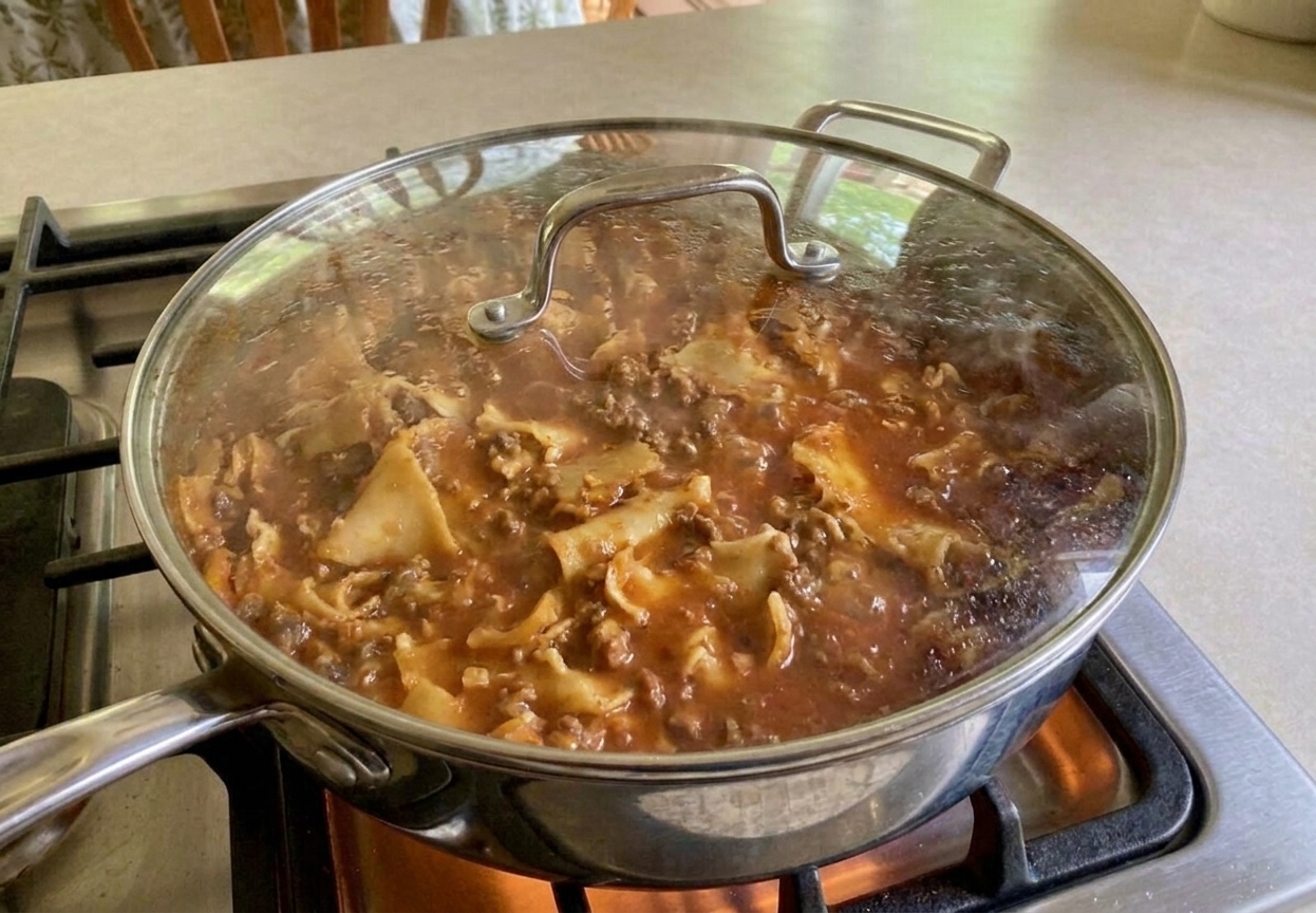 Broken lasagna noodles simmering in a red meat sauce inside a covered skillet with steam rising