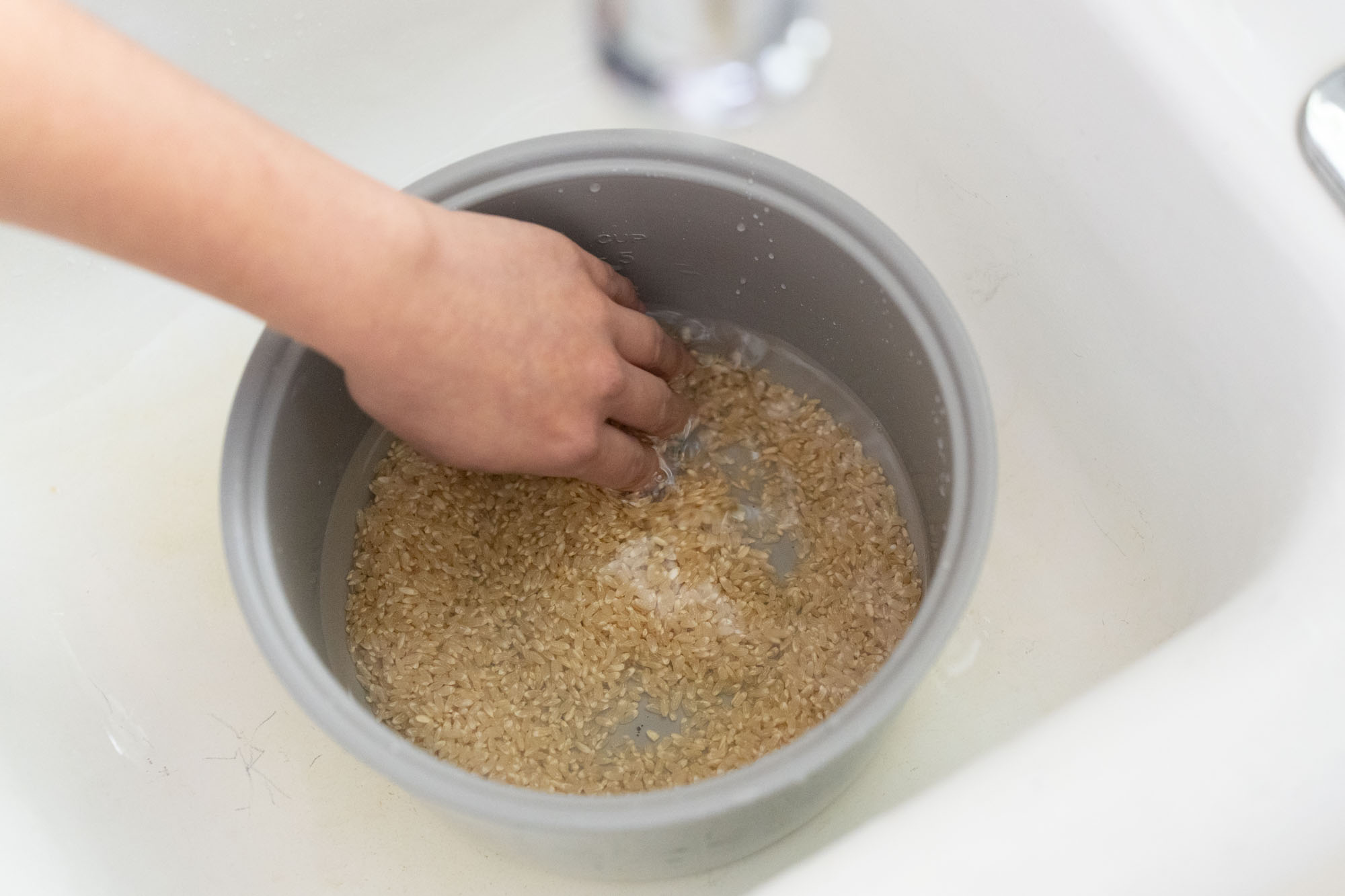 Brown rice being rinsed under running water in a fine-mesh strainer set over a bowl in a home kitchen sink