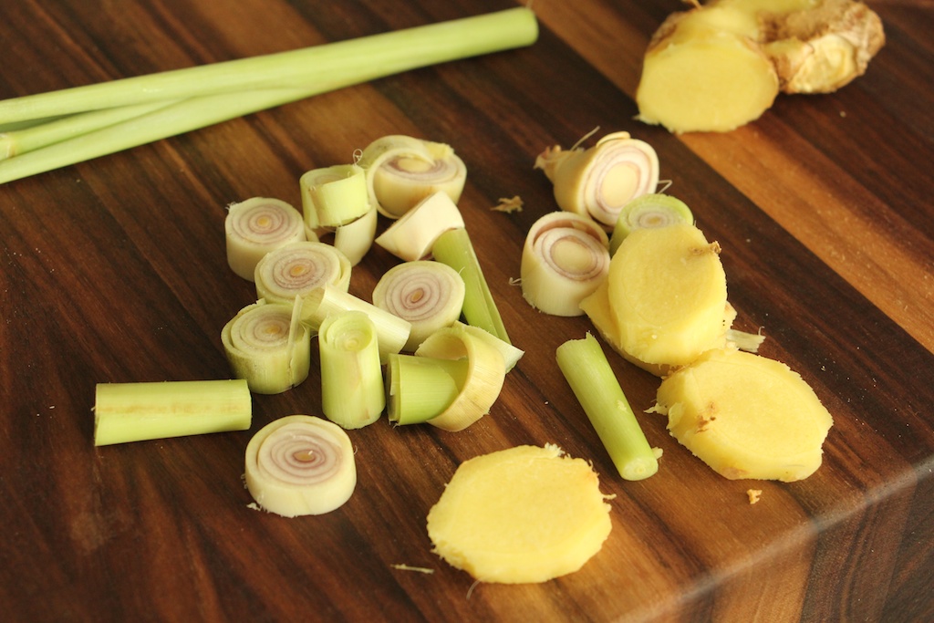 Bruised lemongrass stalks and sliced ginger on a wooden cutting board next to a chef's knife