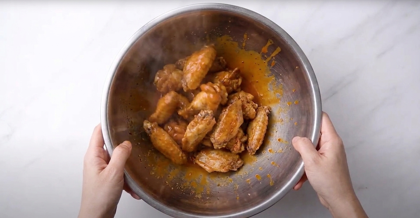 Buffalo wings being tossed in a large stainless steel bowl as warm sauce coats the wings