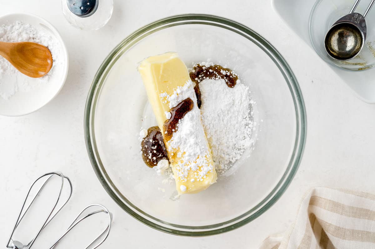 Butter, powdered sugar, vanilla extract, heavy cream, and salt measured out on a clean kitchen counter with mixing bowls