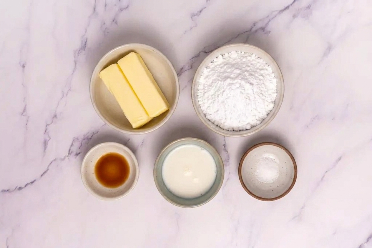 Butter, powdered sugar, vanilla extract, milk, and salt measured out in small bowls on a clean kitchen counter under soft natural light