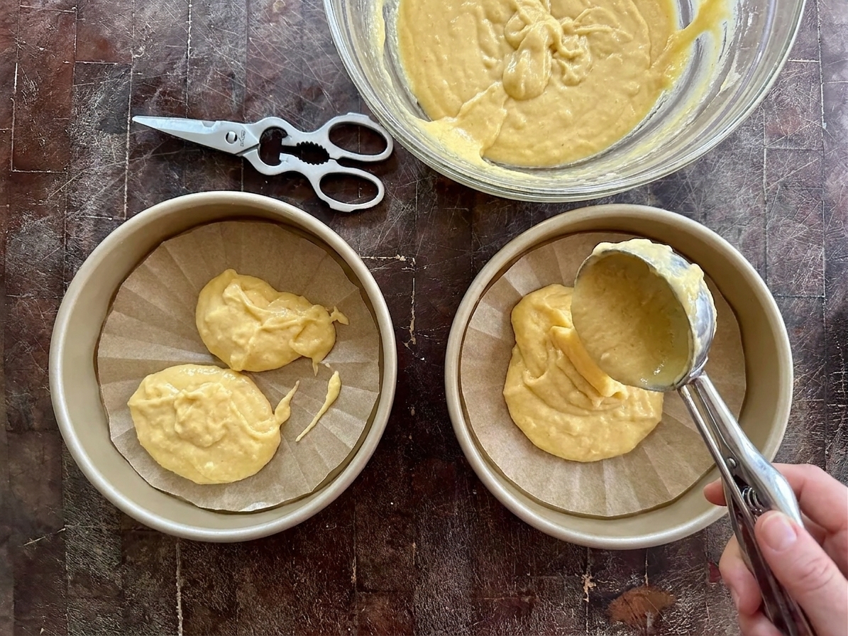 Cake batter being divided evenly between two round pans on a kitchen counter
