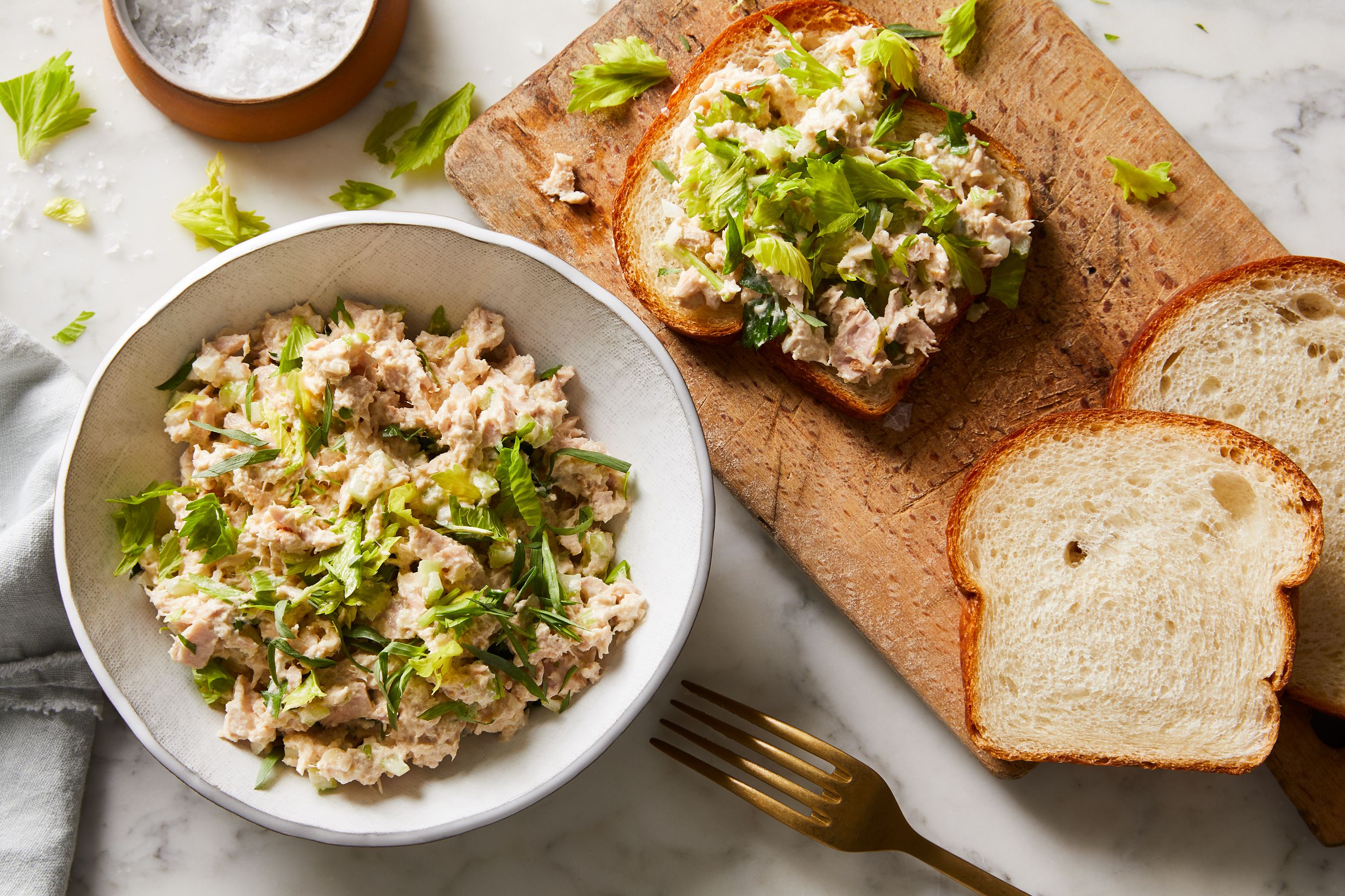 Canned tuna, mayonnaise, Dijon mustard, lemon, celery, pickles, salt, pepper, and bread arranged on a clean kitchen counter