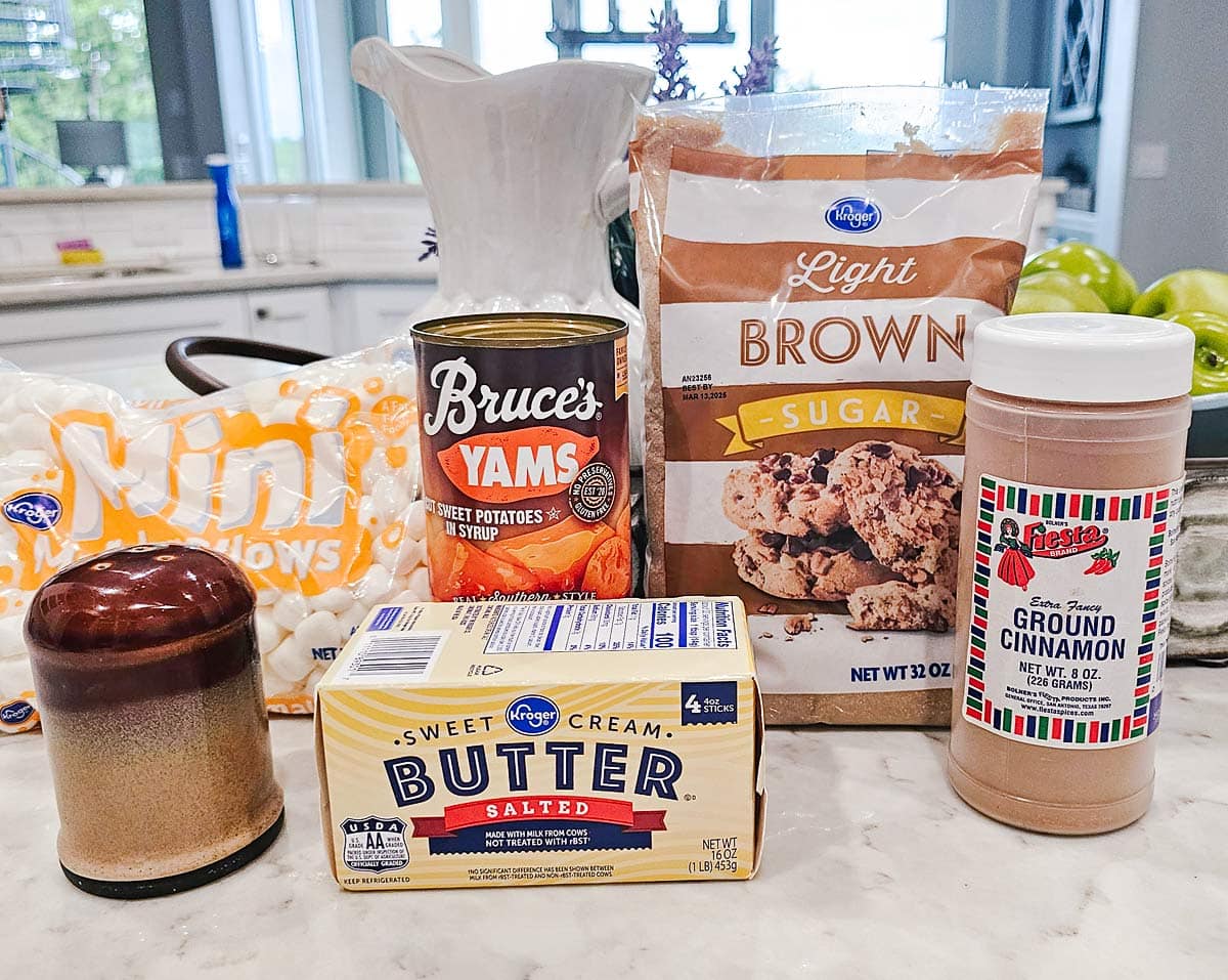 Canned yams, brown sugar, butter, cinnamon, nutmeg, vanilla, and pecans arranged on a light kitchen counter with measuring spoons