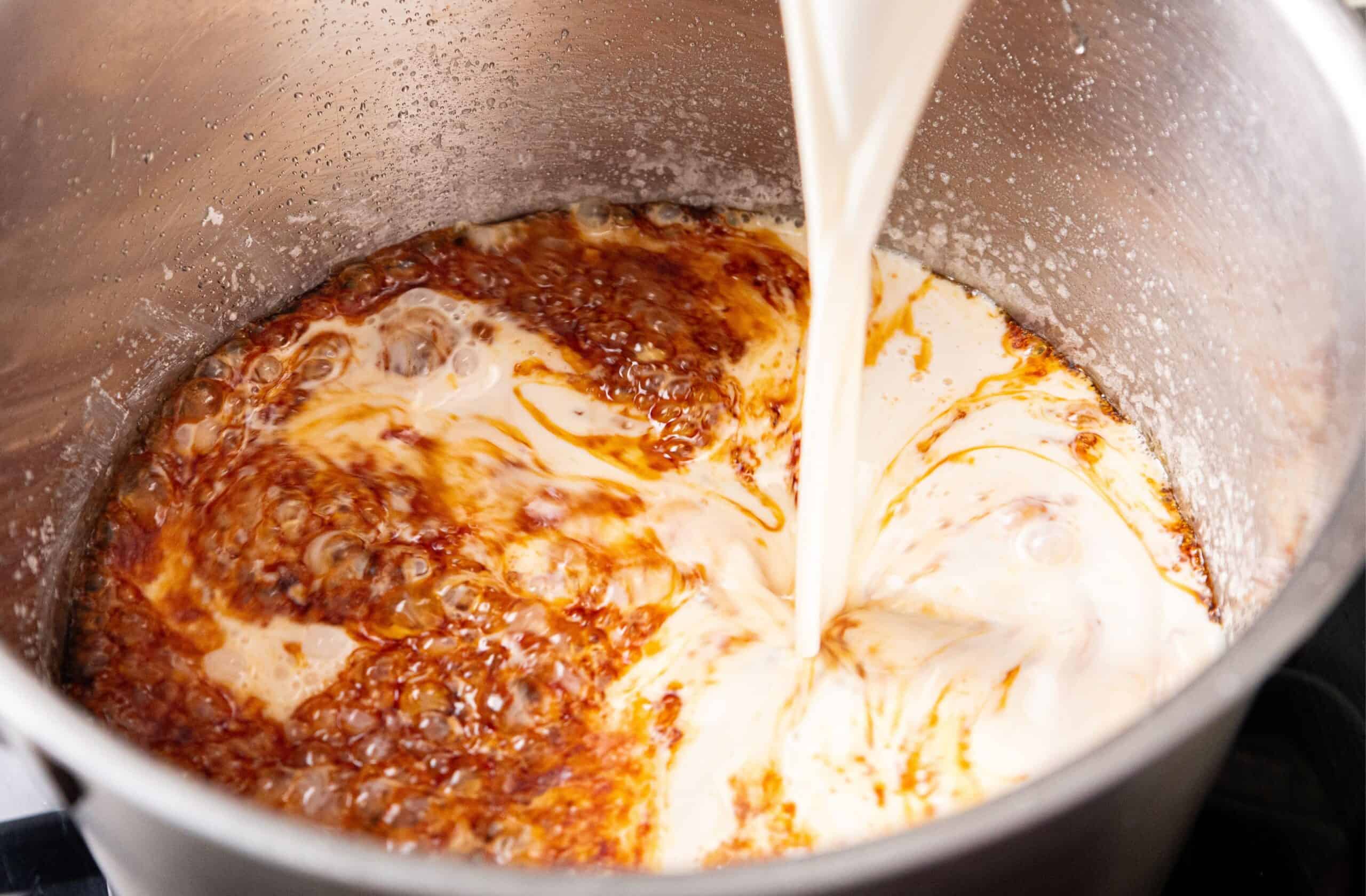 Caramel sauce actively bubbling in a stainless steel saucepan on a stovetop while a whisk is held above the pan