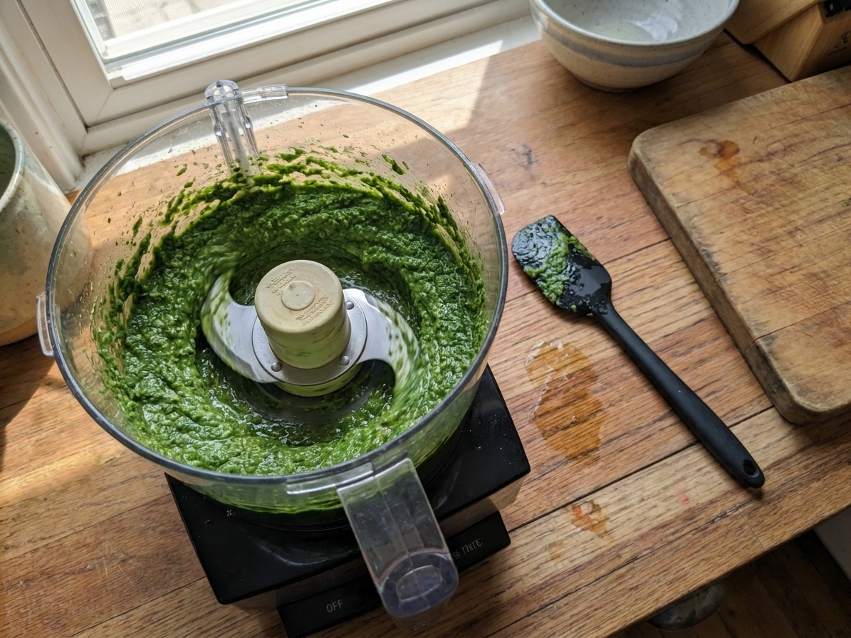 Carrot top pesto being blended in a food processor on a kitchen counter, with a spatula nearby
