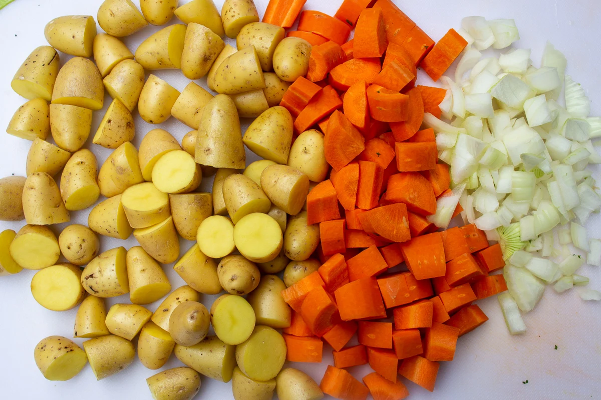 Carrots, baby potatoes, and sliced onion prepped in separate piles on a wooden cutting board