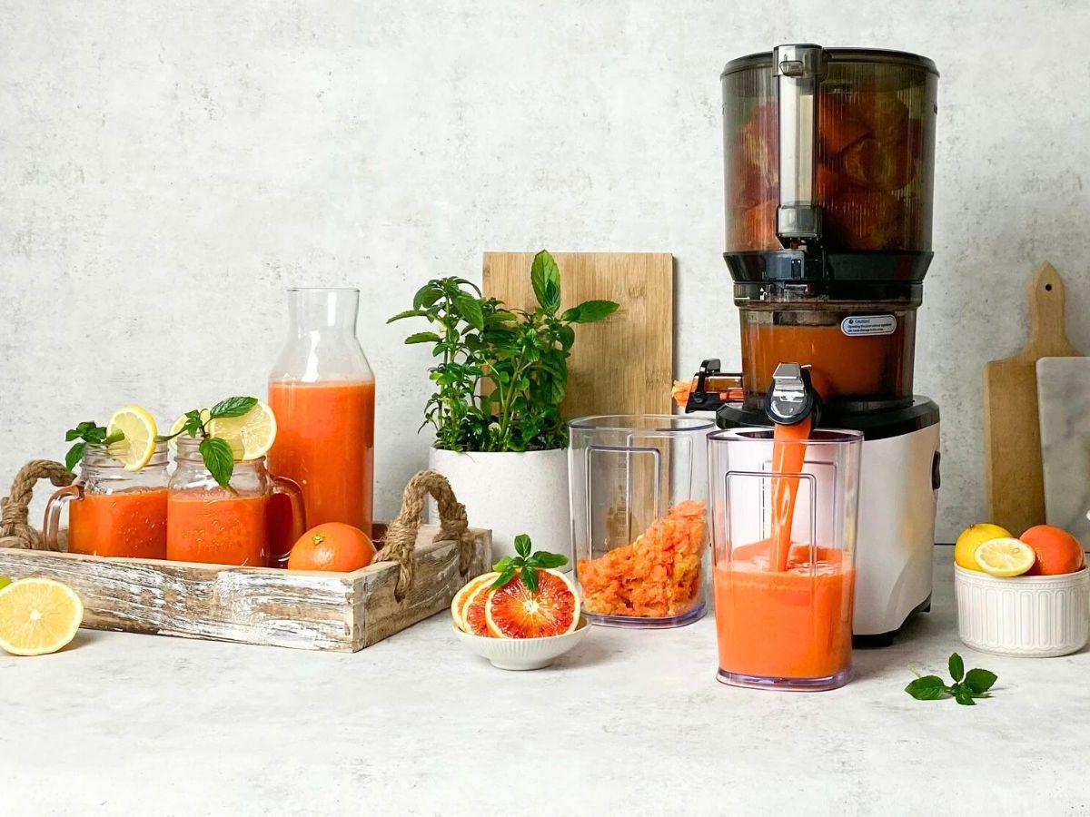 Carrots being fed into a countertop juicer on a bright kitchen counter with a pitcher collecting vivid orange juice