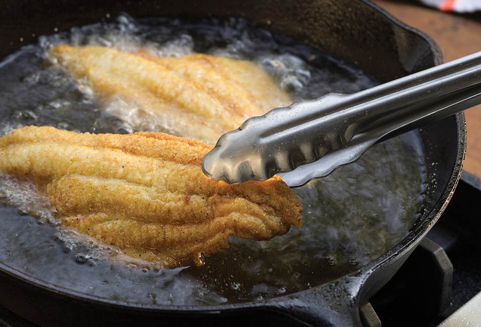 Catfish fillets frying in bubbling oil in a cast iron skillet on a stovetop with tongs nearby