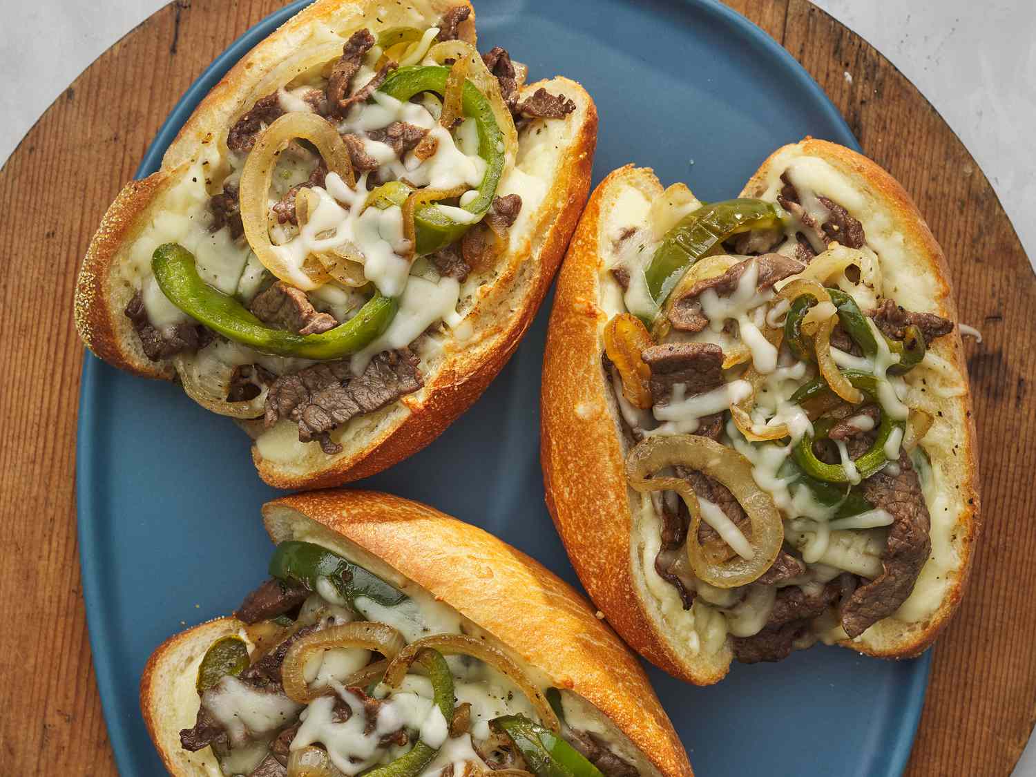 Cheesesteak ingredients laid out on a bright kitchen counter including shaved steak, hoagie rolls, onions, provolone slices, bell pepper, and seasonings in small bowls