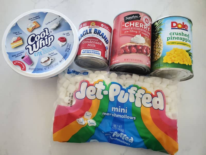Cherry pie filling, mini marshmallows, crushed pineapple, sour cream, and whipped topping arranged on a clean kitchen counter