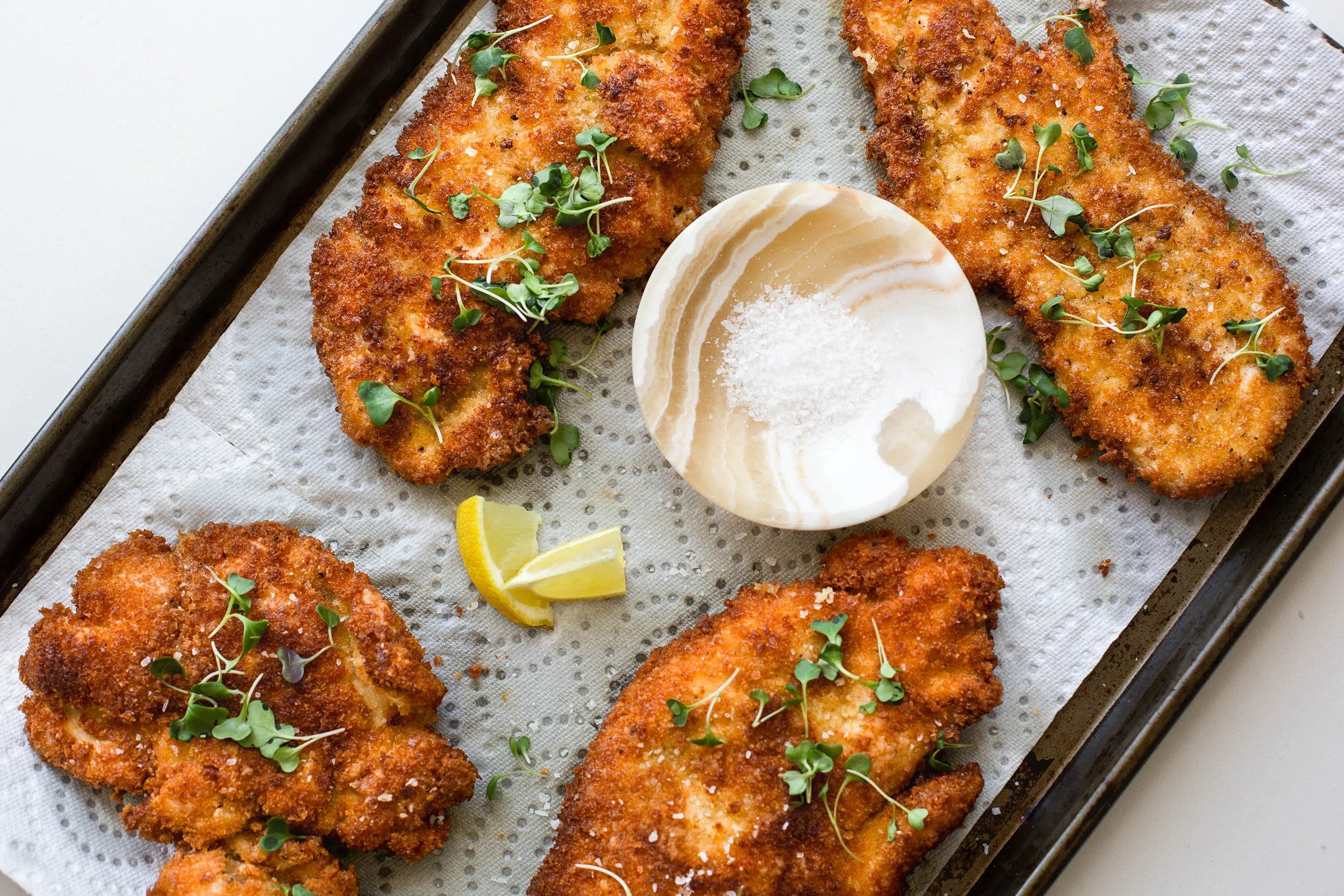 Chicken breast cutlets on a cutting board being seasoned with salt, pepper, and Italian seasoning in a home kitchen