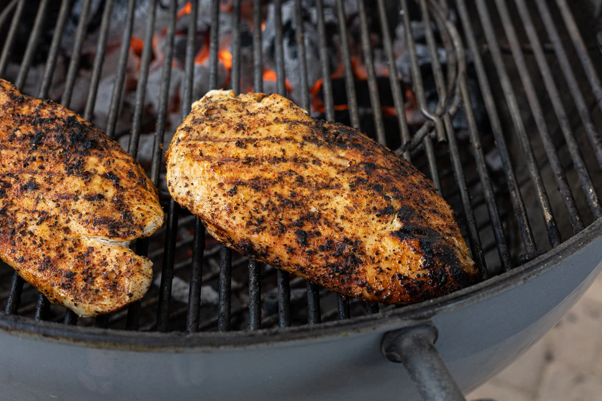 Chicken breasts grilling on hot grates with visible char marks and tongs turning one piece