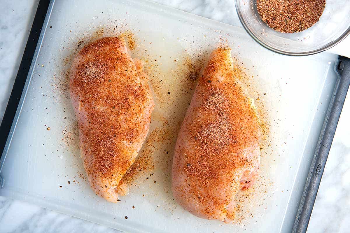 Chicken breasts on a cutting board with small bowls of paprika, garlic powder, salt, pepper, and cumin nearby on a bright kitchen counter