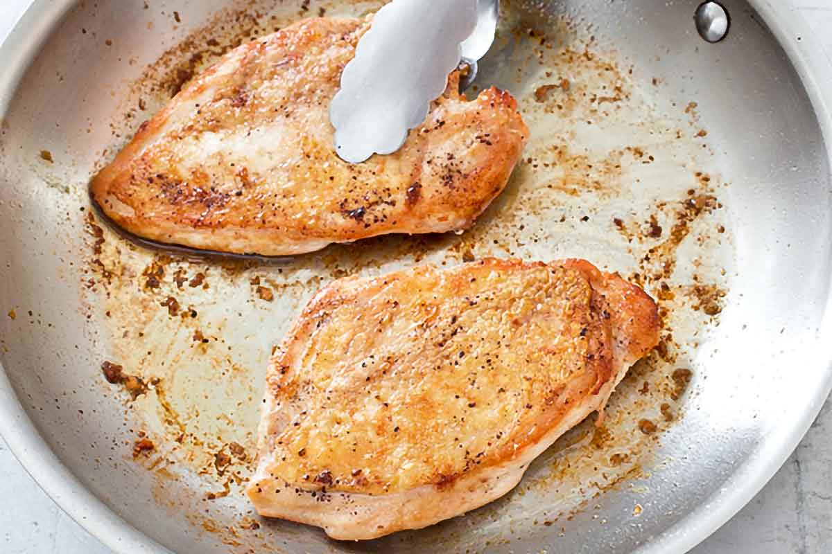 Chicken breasts searing in a cast iron skillet on a stovetop with browned edges forming