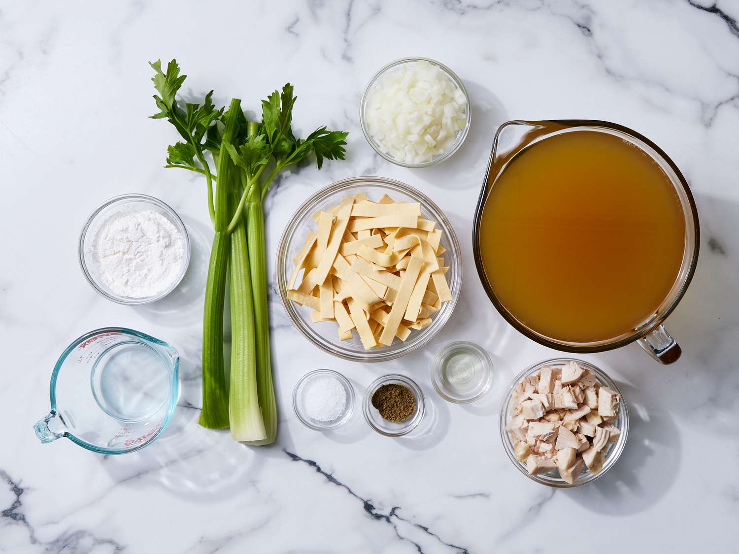 Chicken broth, carrot, celery, onion, garlic, ginger, soy sauce, mushrooms, scallions, and fried onions arranged on a light stone kitchen counter