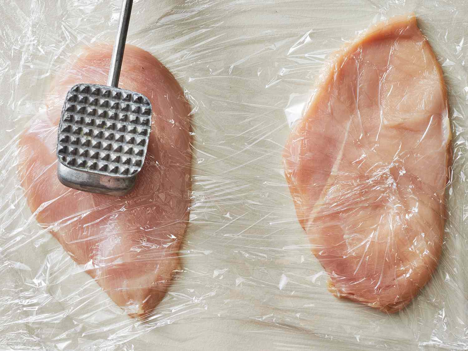 Chicken cutlets being pounded under plastic wrap with a meat mallet on a cutting board in a home kitchen
