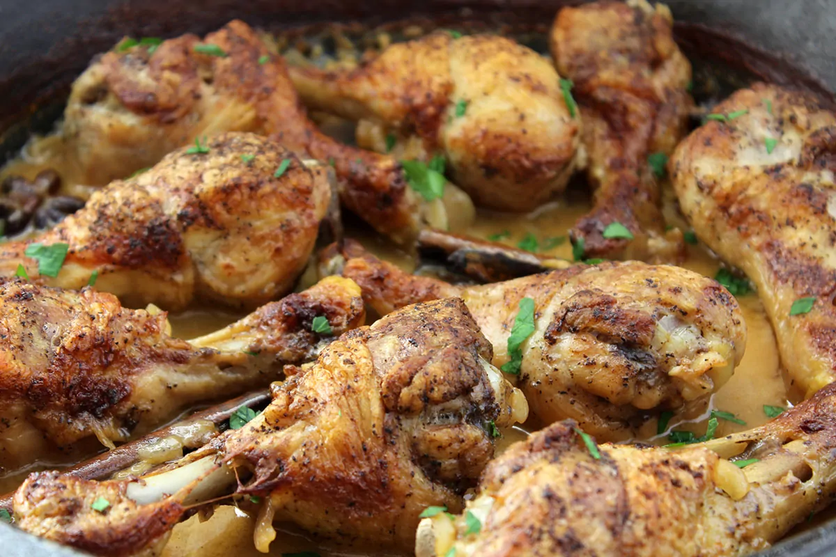 Chicken drumsticks browning in a hot skillet on a stovetop with tongs nearby, warm kitchen lighting