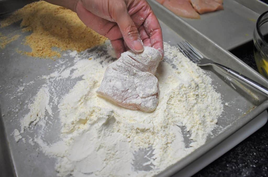 Chicken pieces being coated in seasoned flour in a shallow dish on a kitchen counter, with craggy bits of flour clinging to the surface