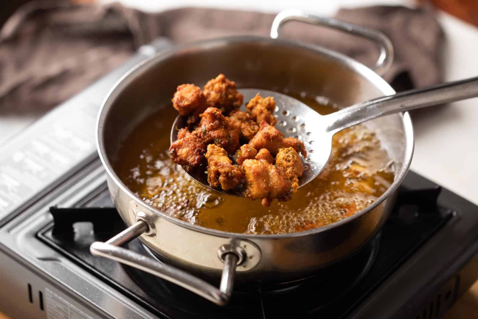 Chicken pieces frying in bubbling oil in a deep skillet on a stovetop, with a wire rack nearby for draining