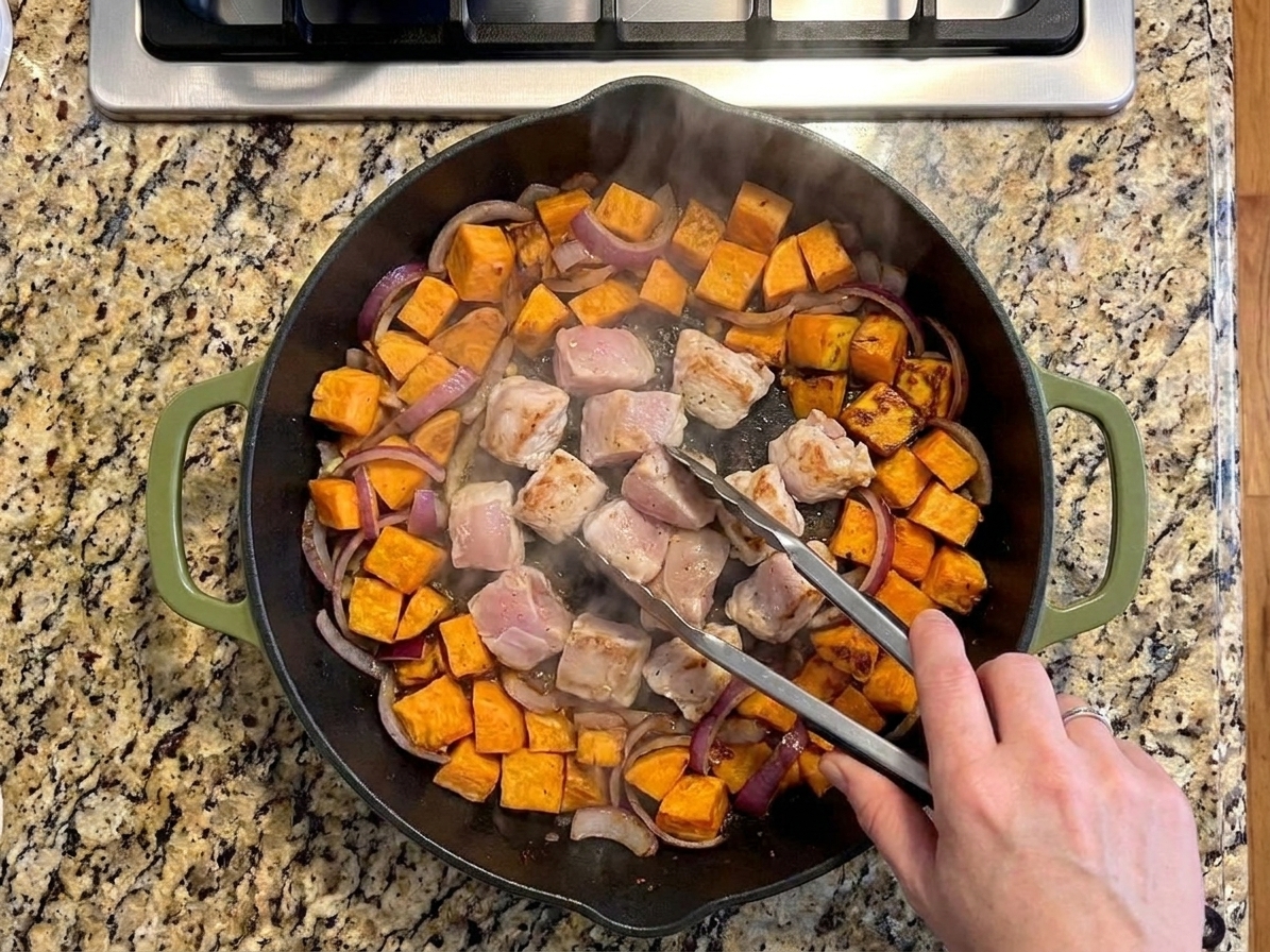 Chicken pieces searing in the center of a cast iron skillet with sweet potatoes and red onions pushed to the edges, steam rising over a stovetop