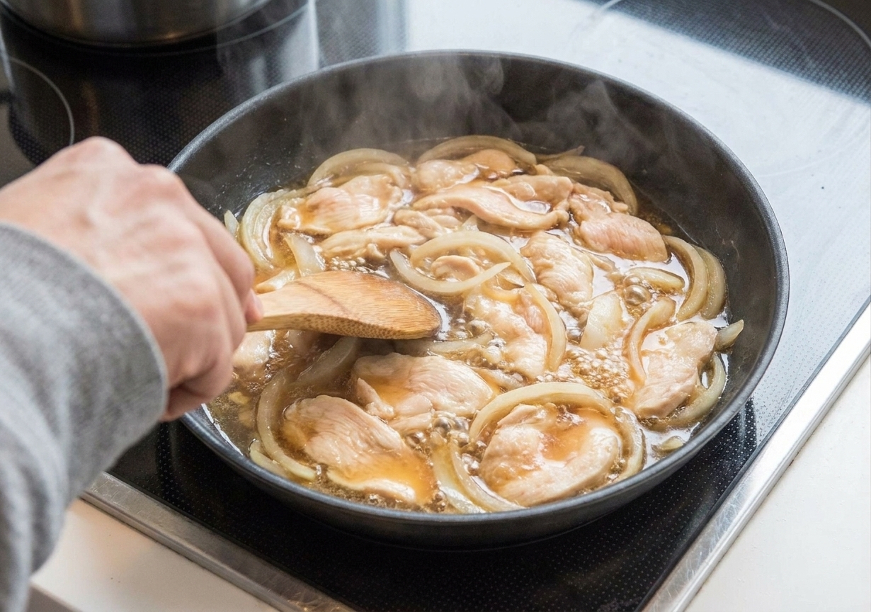 Chicken slices and onions simmering in a shallow soy-mirin-dashi broth in a skillet on the stove