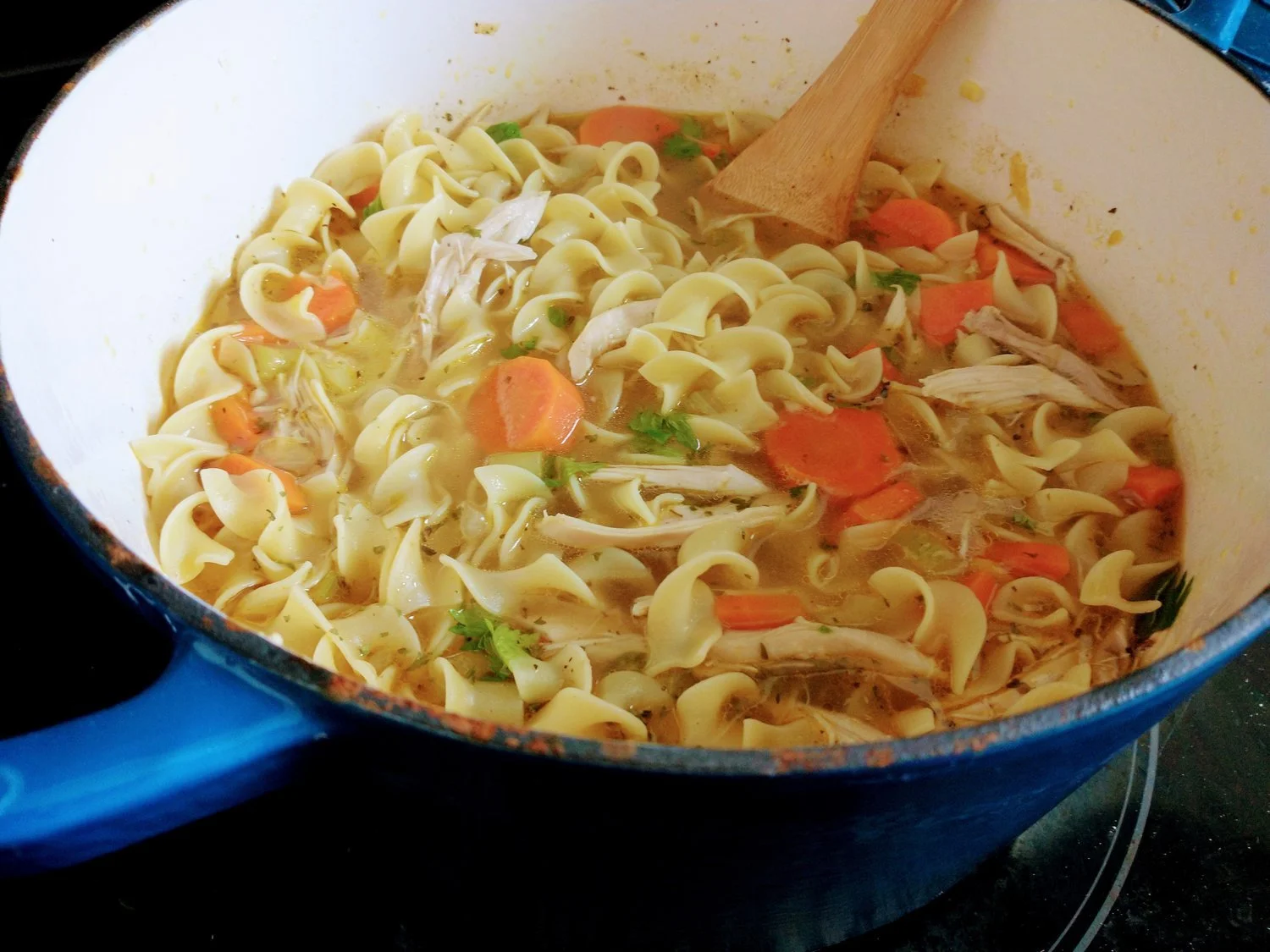 Chicken soup simmering in a Dutch oven on a stovetop with visible carrots, celery, noodles, and shredded chicken