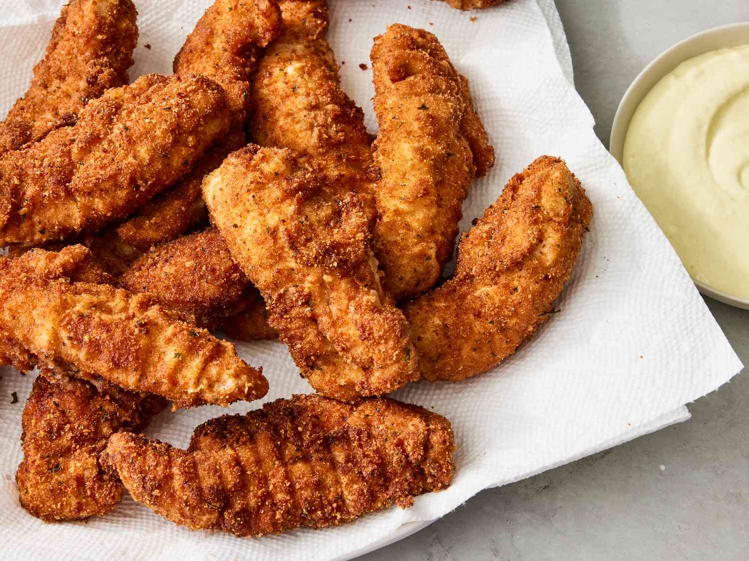 Chicken strips being dipped into egg and pressed into breadcrumbs over three shallow bowls on a kitchen counter with warm, consistent lighting