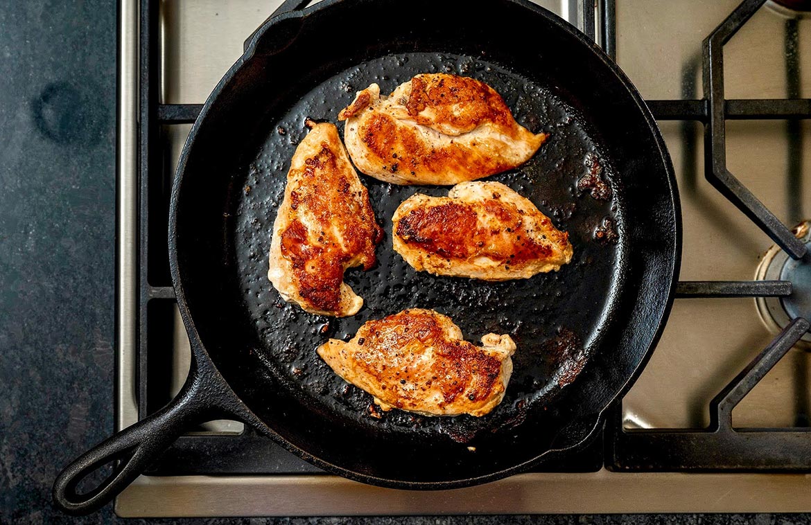 Chicken thigh pieces sizzling in a large skillet with browned edges forming, viewed from above on a stovetop