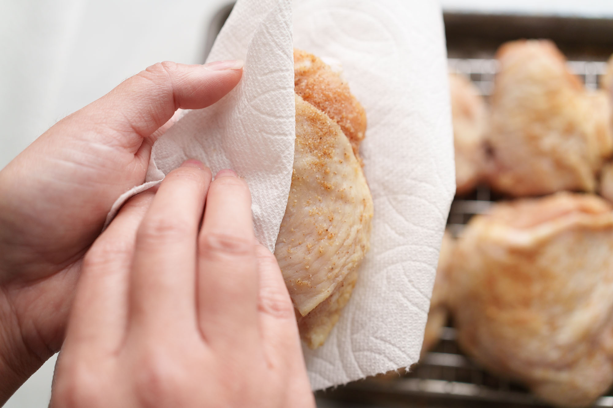 Chicken thighs being patted very dry with paper towels on a cutting board next to a cast iron skillet on the stove