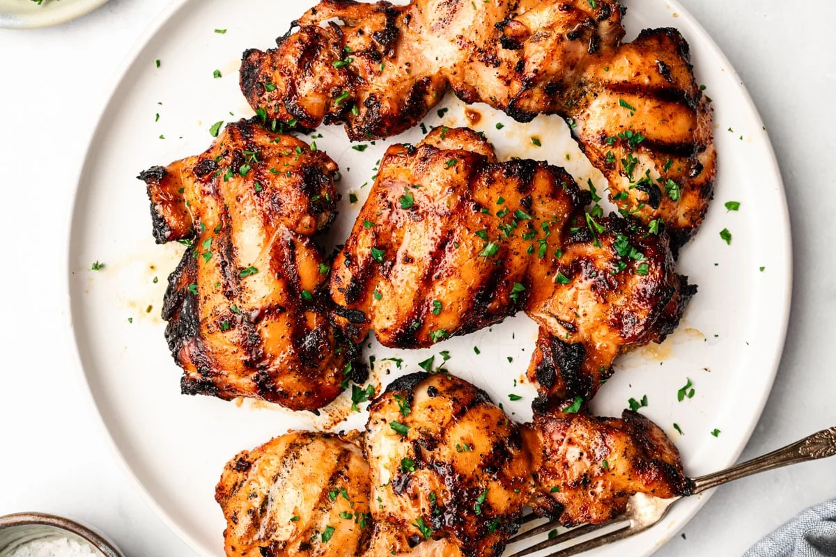 Chicken thighs sizzling on a grill grate with visible char marks and a small flare of heat, outdoor cooking action shot