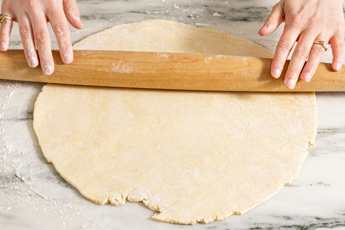 Chilled tart dough being rolled into a rough circle on a lightly floured kitchen counter with a wooden rolling pin