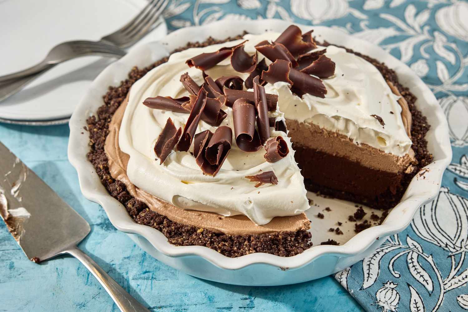 Chocolate cookie crumbs being pressed firmly into a 9-inch pie dish using the bottom of a measuring cup on a clean kitchen counter