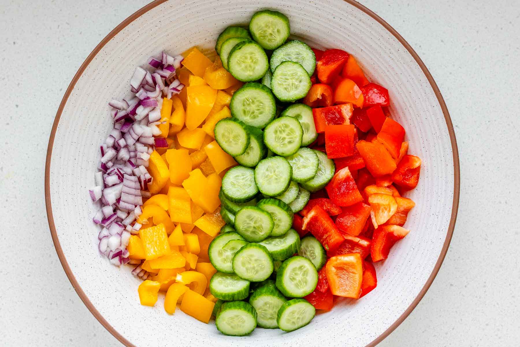 Chopped cucumbers, diced onion, and diced green bell pepper in a glass bowl with salt being sprinkled over the top on a kitchen counter