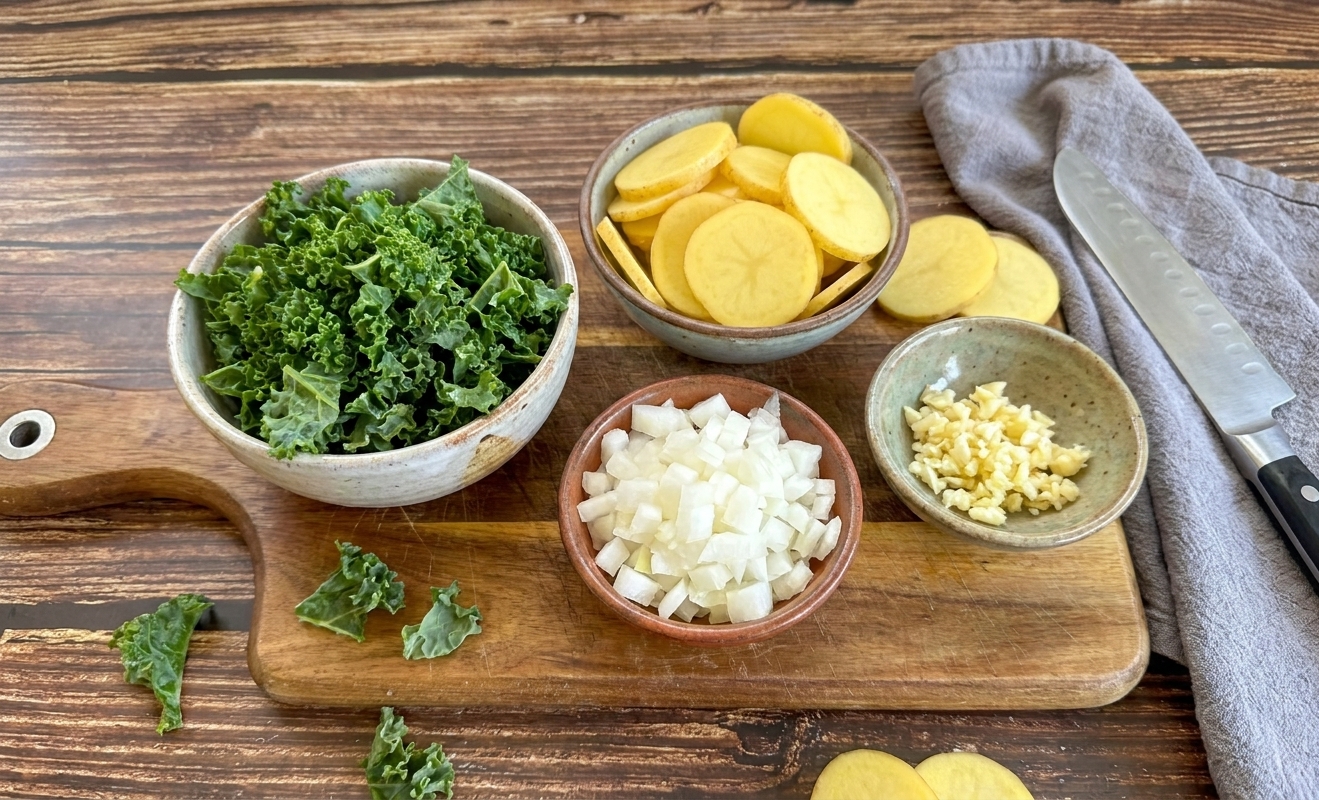 Chopped kale, sliced potatoes, diced onion, and minced garlic arranged in small bowls on a kitchen counter, ready for cooking