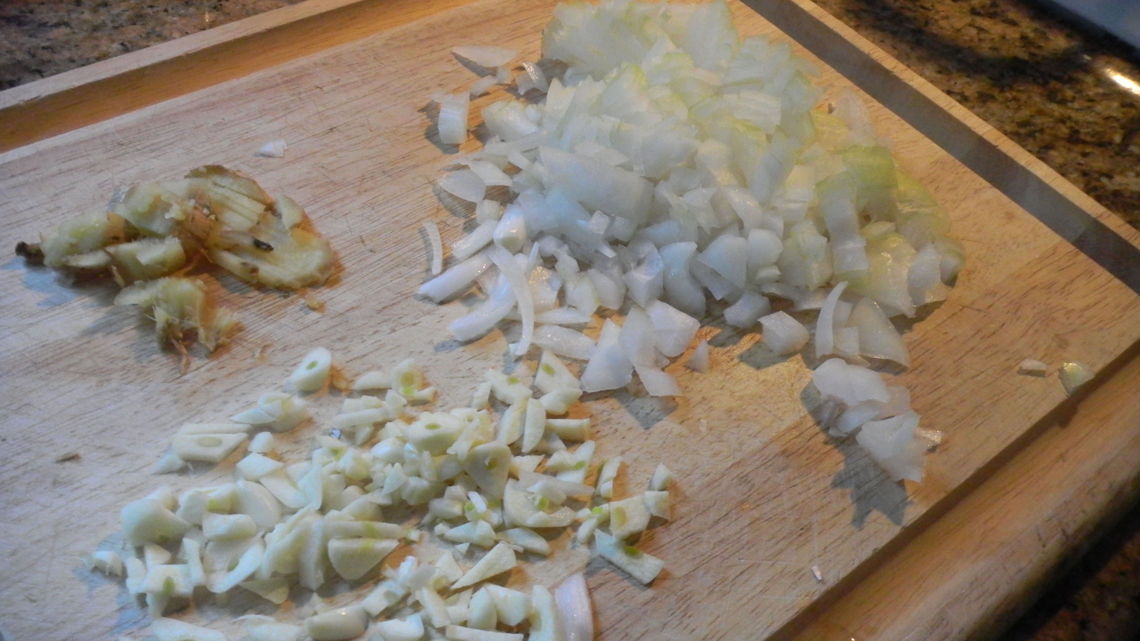 Chopped onion, minced garlic, and grated ginger in small piles on a wooden cutting board beside a chef's knife