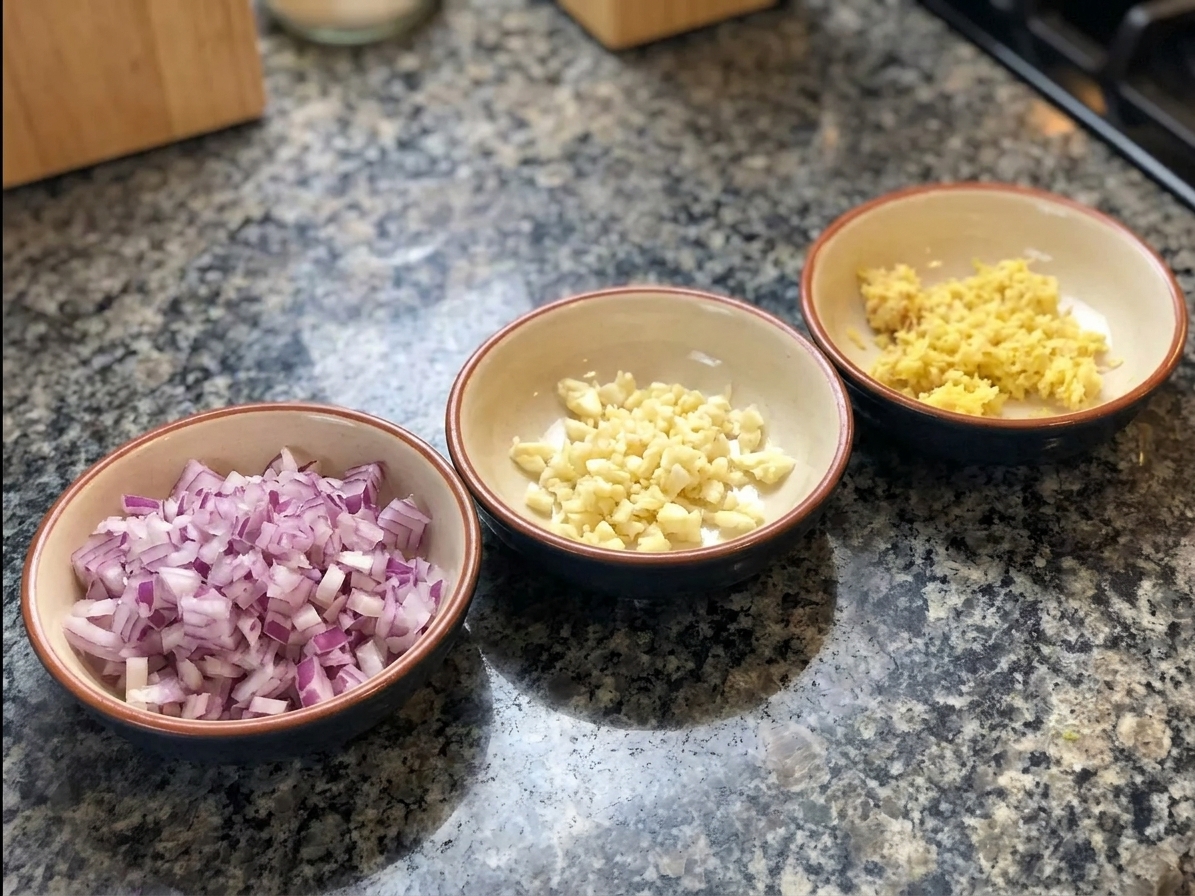 Chopped onion, minced garlic, and grated ginger prepped in small bowls on a kitchen counter