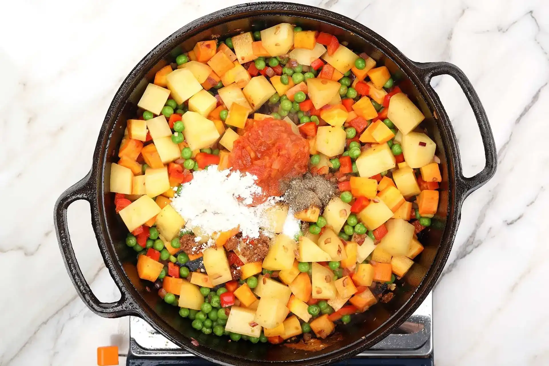 Chopped onions, minced garlic, potato chunks, sliced carrots, and sliced red bell pepper arranged in separate piles on a wooden cutting board on a kitchen counter