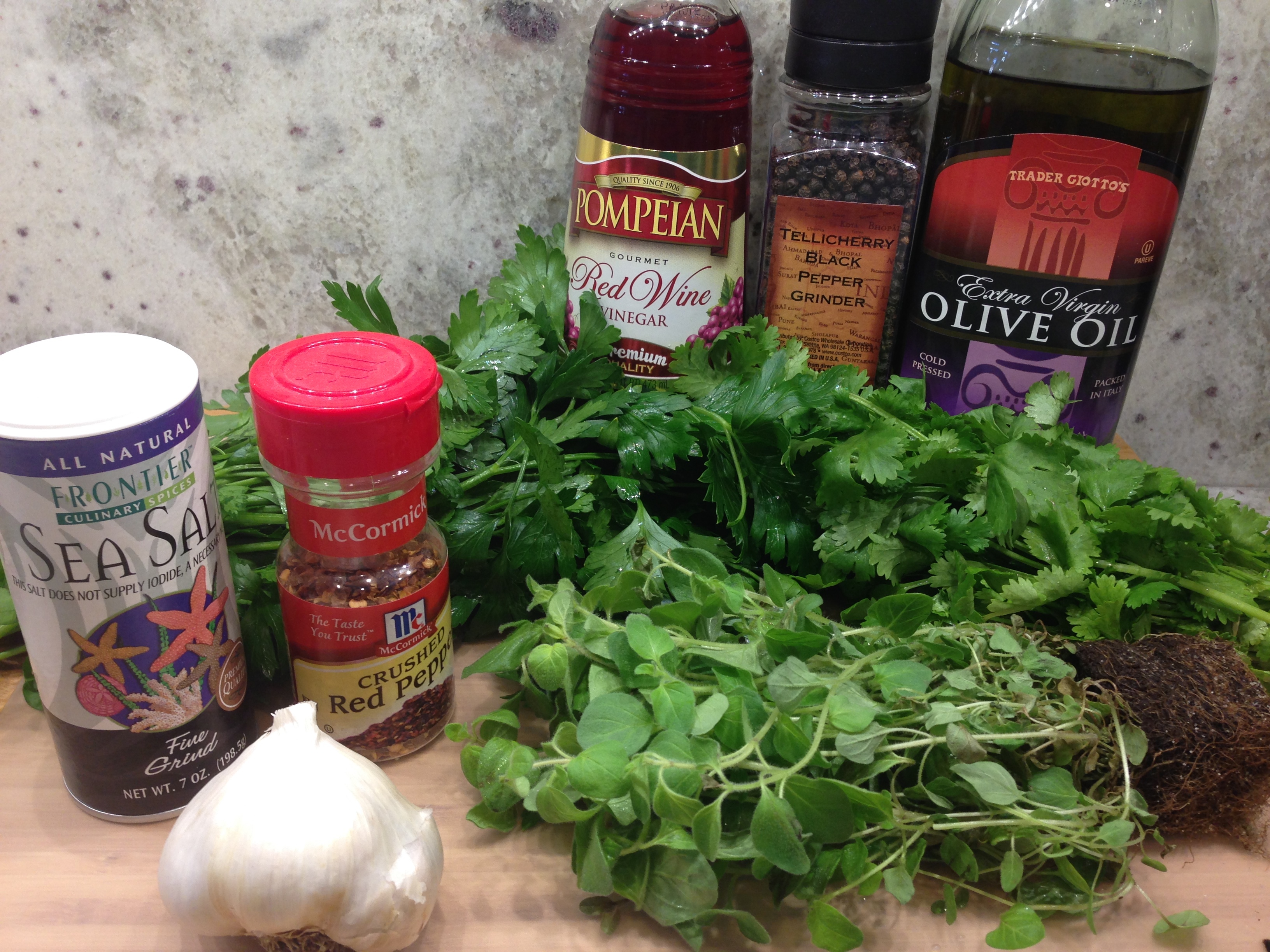 Chopped parsley, chopped cilantro, minced garlic, red wine vinegar, olive oil, salt, pepper, and red pepper flakes arranged around a small mixing bowl on a clean kitchen counter