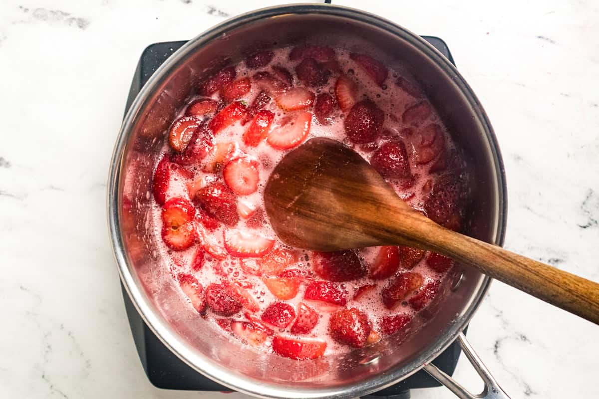 Chopped strawberries simmering in a small saucepan on a stovetop, turning glossy and jammy as they reduce