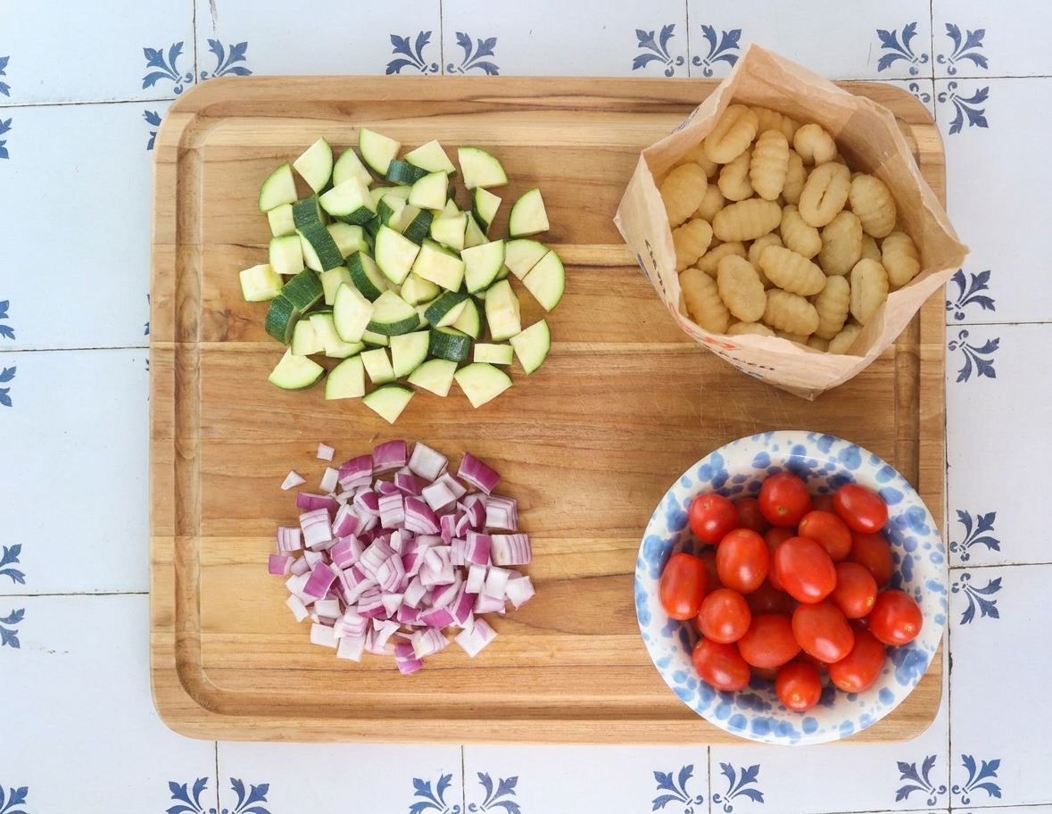 Chopped zucchini and red onion on a cutting board next to a bag of potato gnocchi and a bowl of cherry tomatoes on a bright kitchen counter