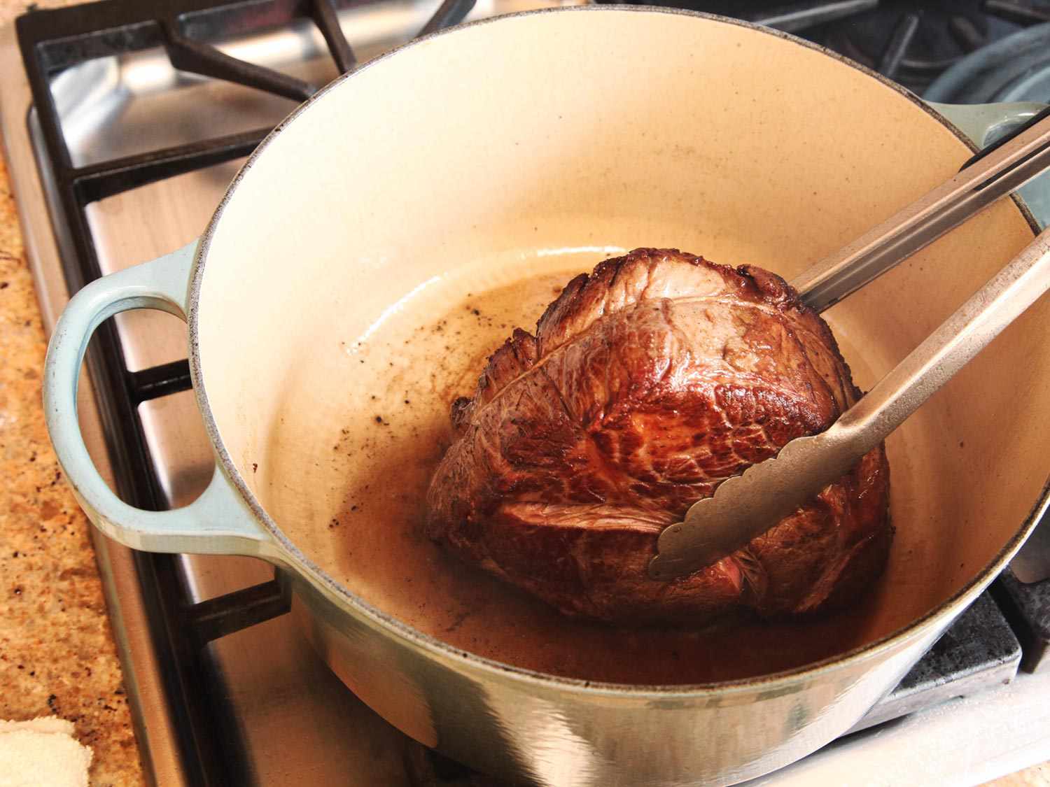 Chunks of beef browning in a Dutch oven on a stovetop with tongs turning a piece to show a deep brown crust