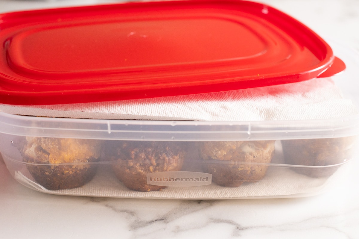 Cinnamon muffins stored in a clear airtight container on a kitchen counter with a paper towel lining