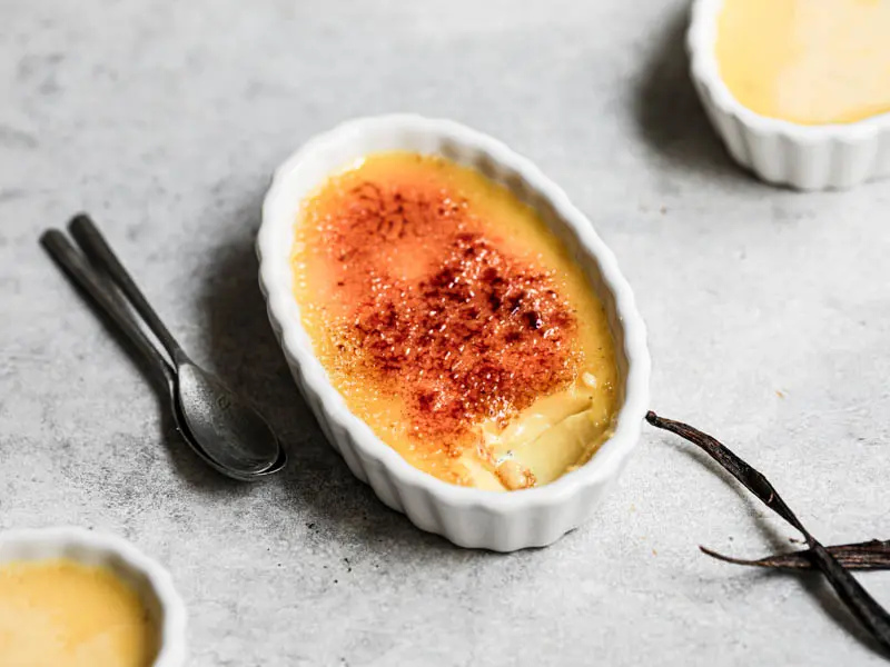 Classic crème brûlée in white ramekins on a rustic kitchen table, with a glossy caramelized sugar crust and a small spoon nearby